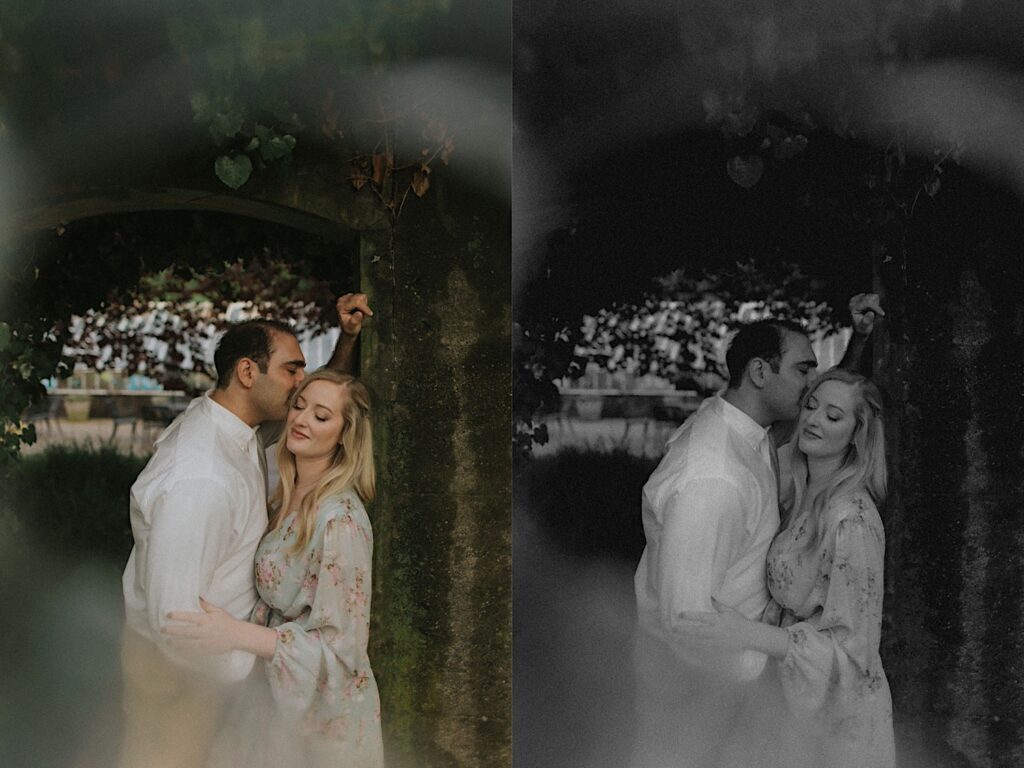 Couple leans up against stone arch as man kisses woman on the head during romantic engagement portraits. 