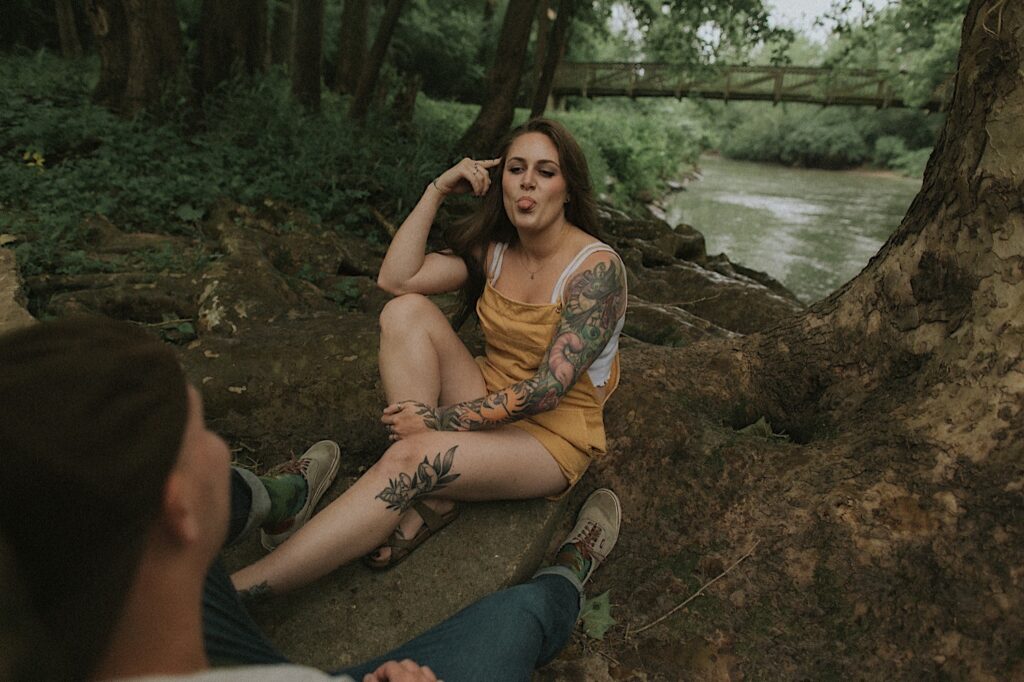 Woman sits underneath a tree in a forested camp ground and sticks her tongue out at her fiance, who's back is facing the camera. 