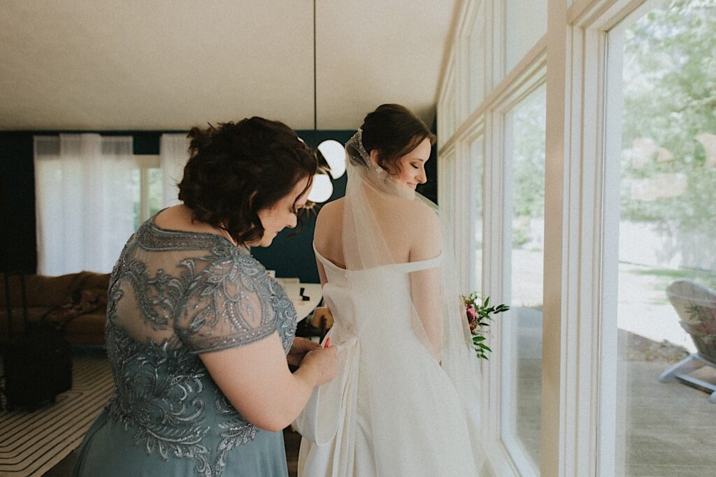 Bride's mom tie's bride's dress into a bow as bride looks behind her shoulder smiling while getting ready before wedding ceremony in Central Illinois. 