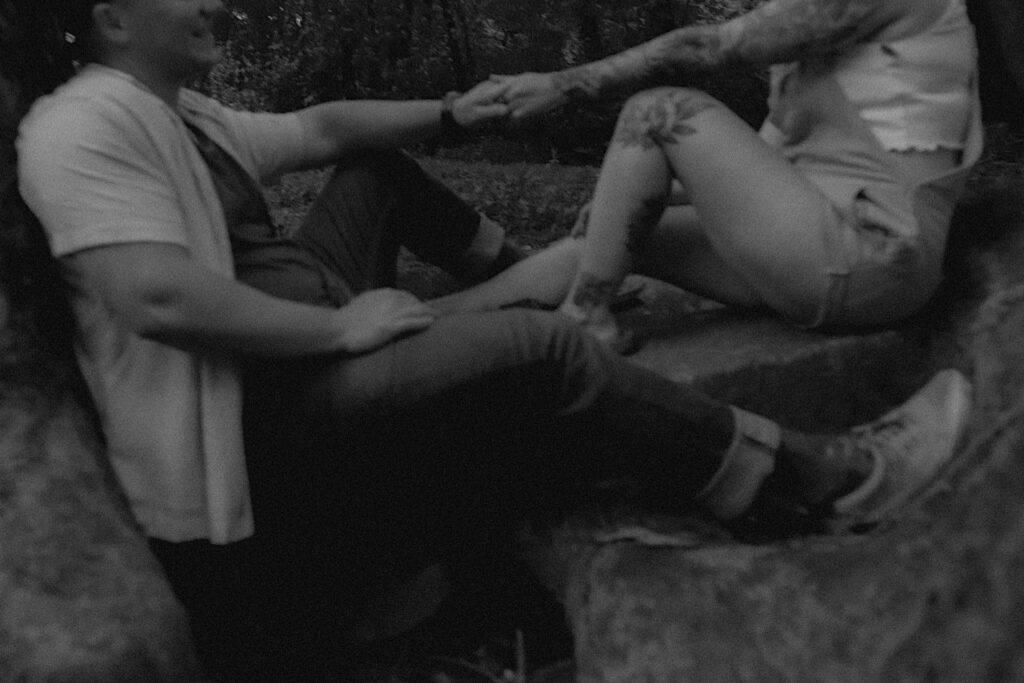 Illinois Engagement Photographer captures the bottom half of a couple holding hands and facing each other as they sit on rocks in a forested park in Lincoln, Illinois. 