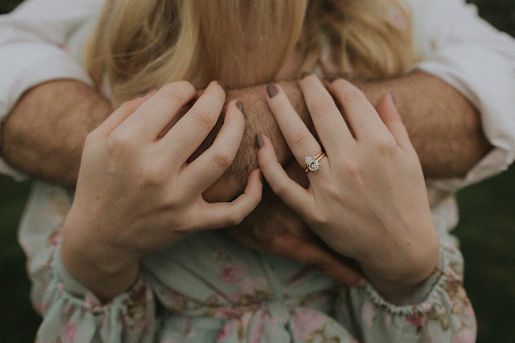 Illinois Engagement photographer captures the details of woman's princess cut engagement ring as she holds her fiance's hand with his arms wrapped around her. 