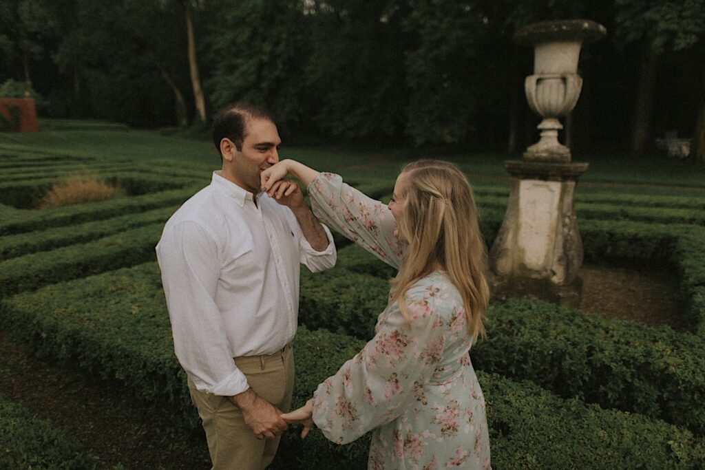 Man holds woman hand and kisses it as they're surrounded by hedge maze. 