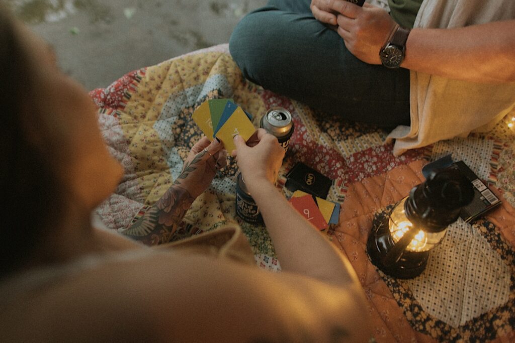 Illinois Engagement photographer captures girl's hands as she holds a hand of uno cards while sitting on a quilt. On the quilt is a lantern, Uno deck and Old Style beers. 