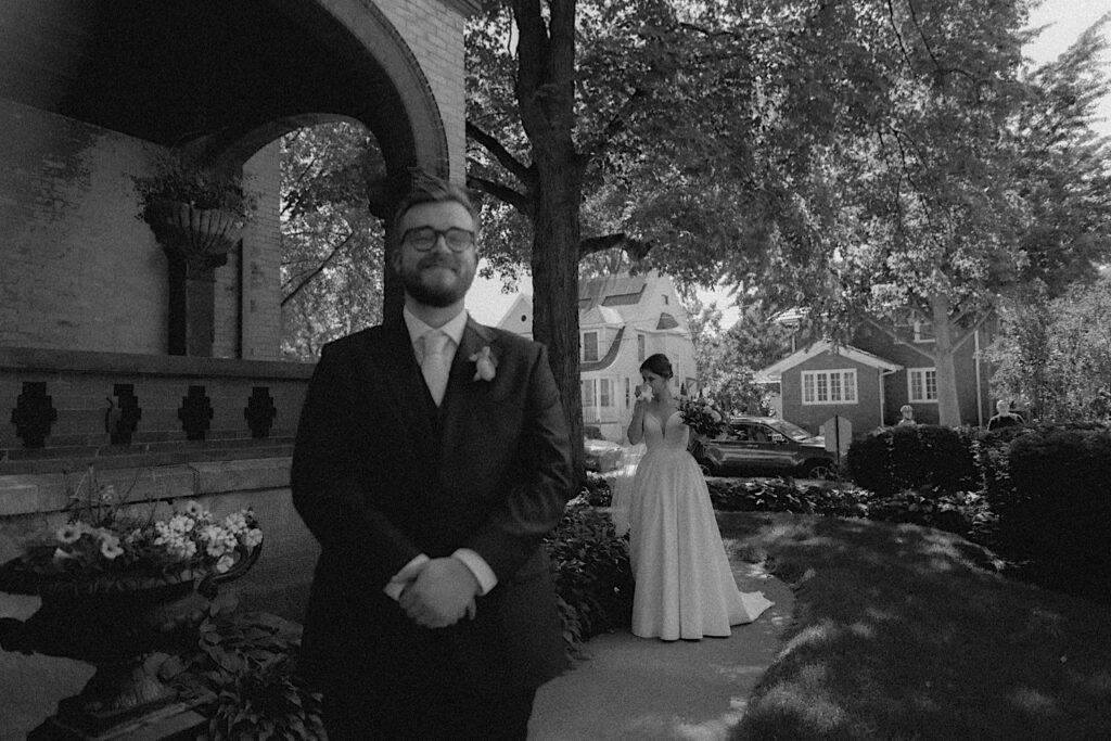 Bride stands behind groom as he waits for first look outside Reality on Monroe in Central Illinois. 