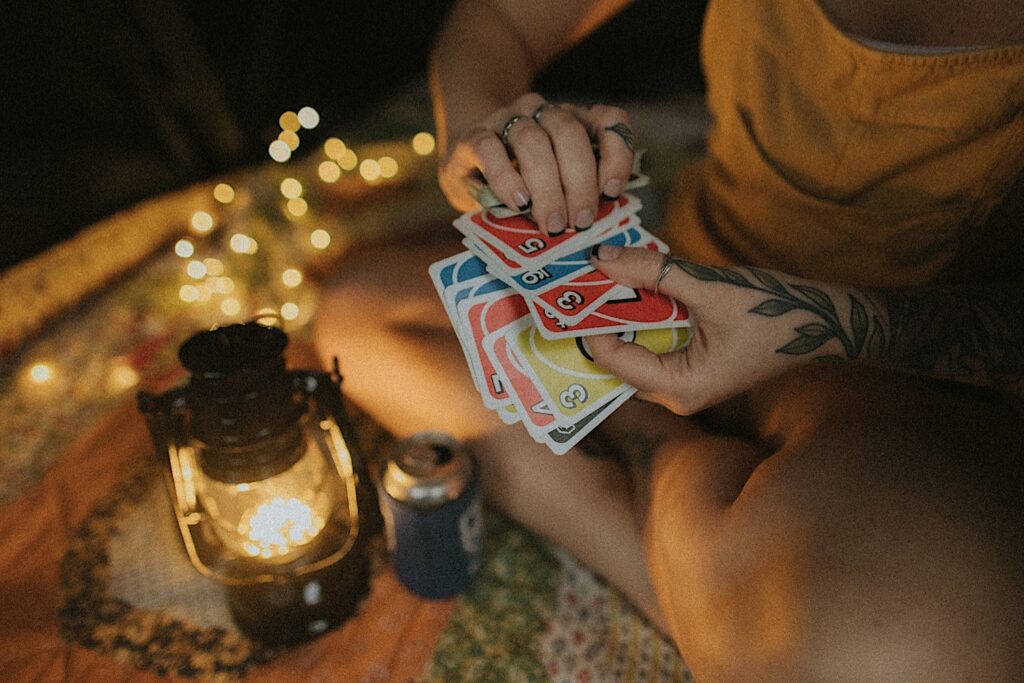 Girl holds onto a deck of Uno cards and shuffles them while sitting on a quilt next to a lantern and a can of beer. 