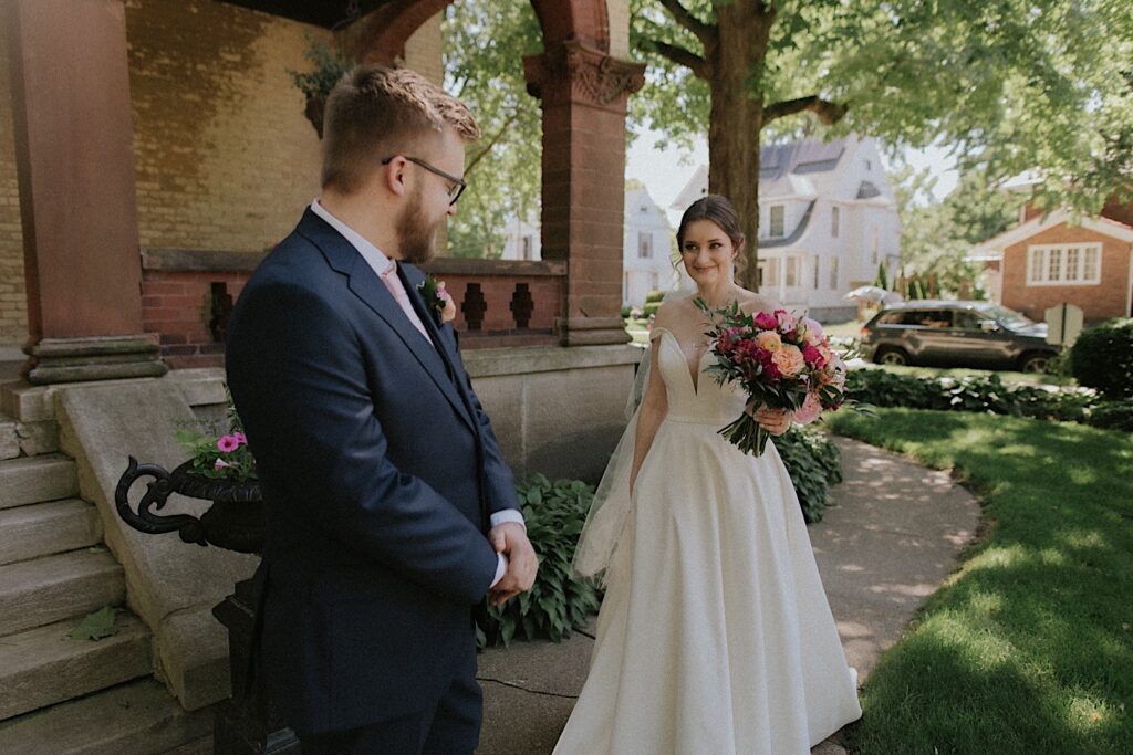 Groom turns around to face bride for first look as she smiles walking towards him holding a brightly pink bouquet outside Reality on Monroe. 