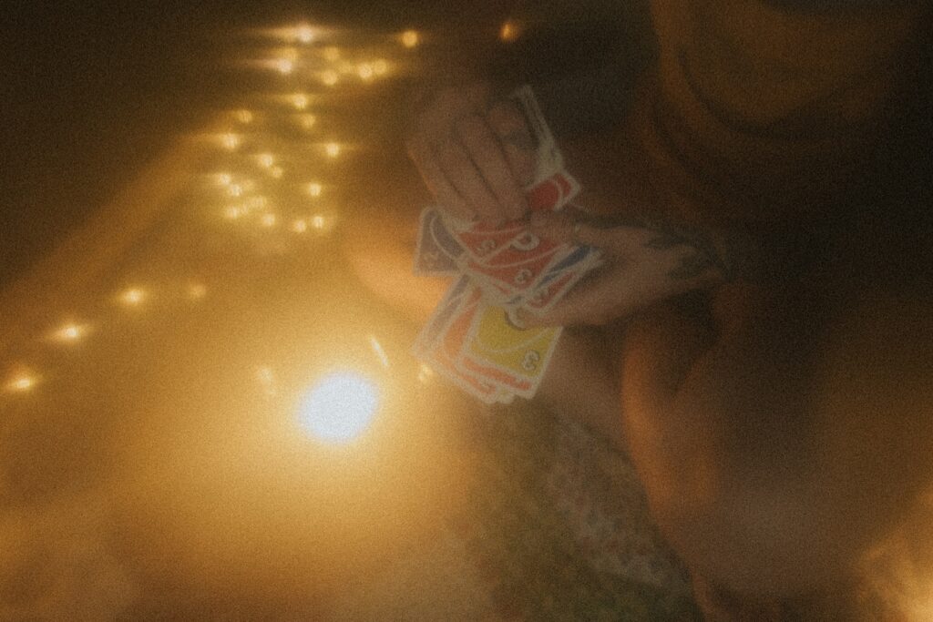 Girl holds onto a deck of Uno cards and shuffles them while sitting on a quilt next to a lantern, string lights and a can of beer. 