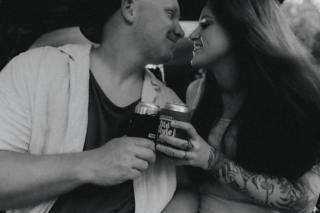 Couple holds cans of Old Style Beer while pressing their faces together for a moody engagement portrait in Lincoln, Illinois. 