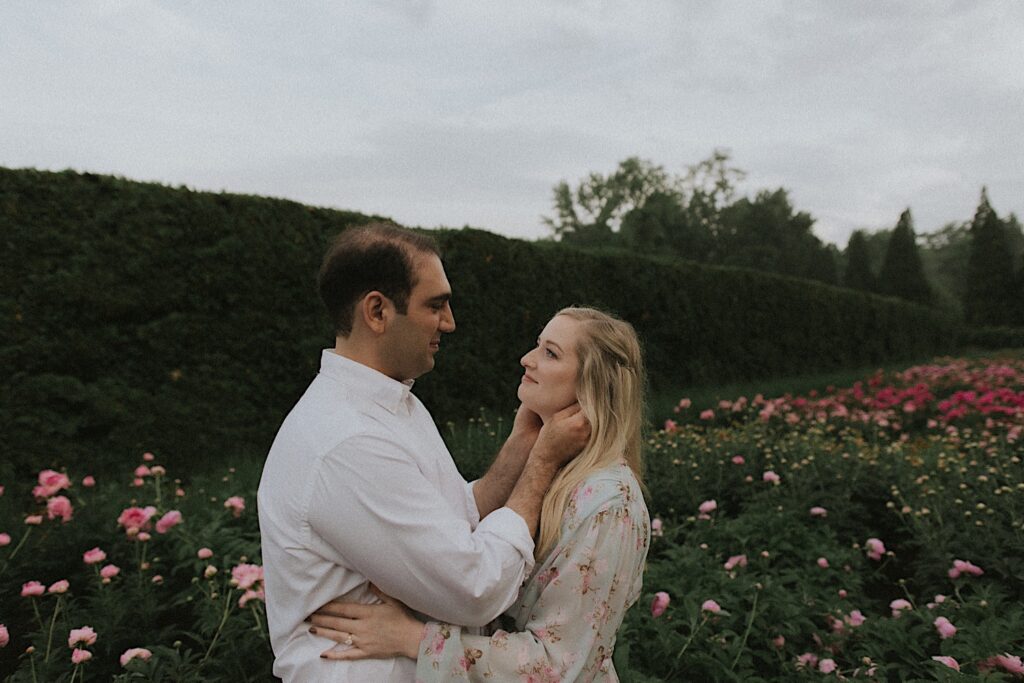 Man holds his fiancee's face as he looks into her eyes as they're surrounded by gorgeous pink flowers in Allerton Park Garden. 