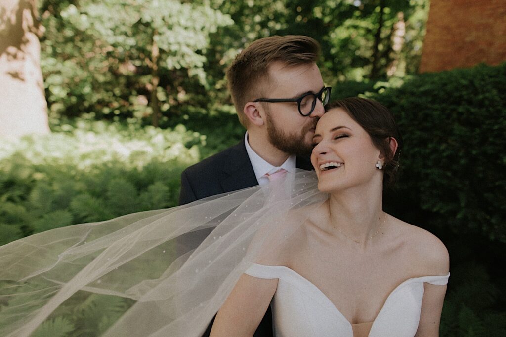 Groom kisses bride's forehead as he hugs her from behind and she laughs for wedding portraits by Central Illinois wedding photographer. 