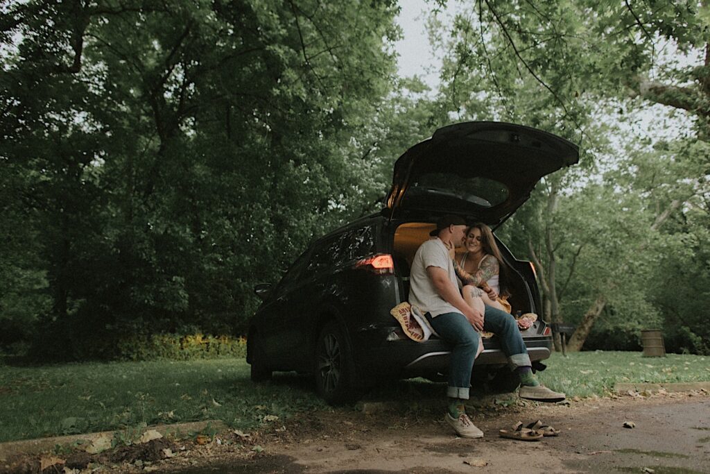 Couple sits in the open trunk of their car on a quilt with their feet hanging off, smiling and looking at each other for unique camping engagement portraits in Lincoln, Illinois. 