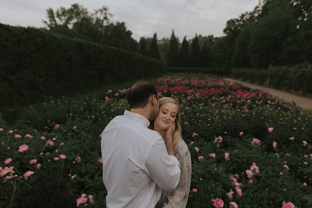 Man kisses woman on the cheek as they stand amongst a garden of pink flowers in Allerton Park. 