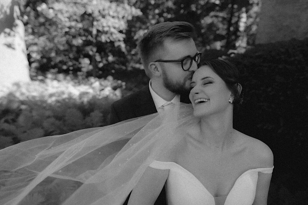 Groom kisses bride's forehead as he hugs her from behind and she laughs for a black and white wedding portrait by Central Illinois wedding photographer. 