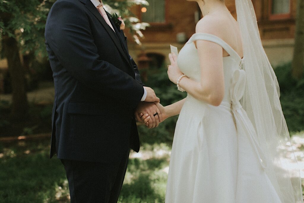 Bride holds grooms hands as she holds her a piece of paper for private vows in front of Reality on Monroe wedding venue. 