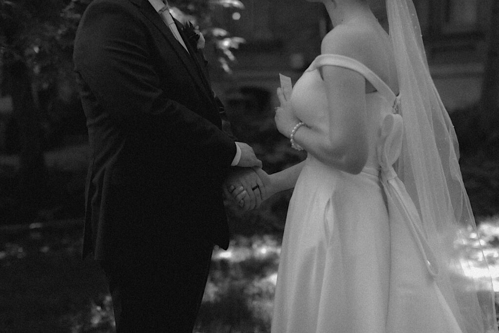 Black and white portrait of bride holding groom's hands as she holds her a piece of paper for private vows in front of Reality on Monroe wedding venue. 