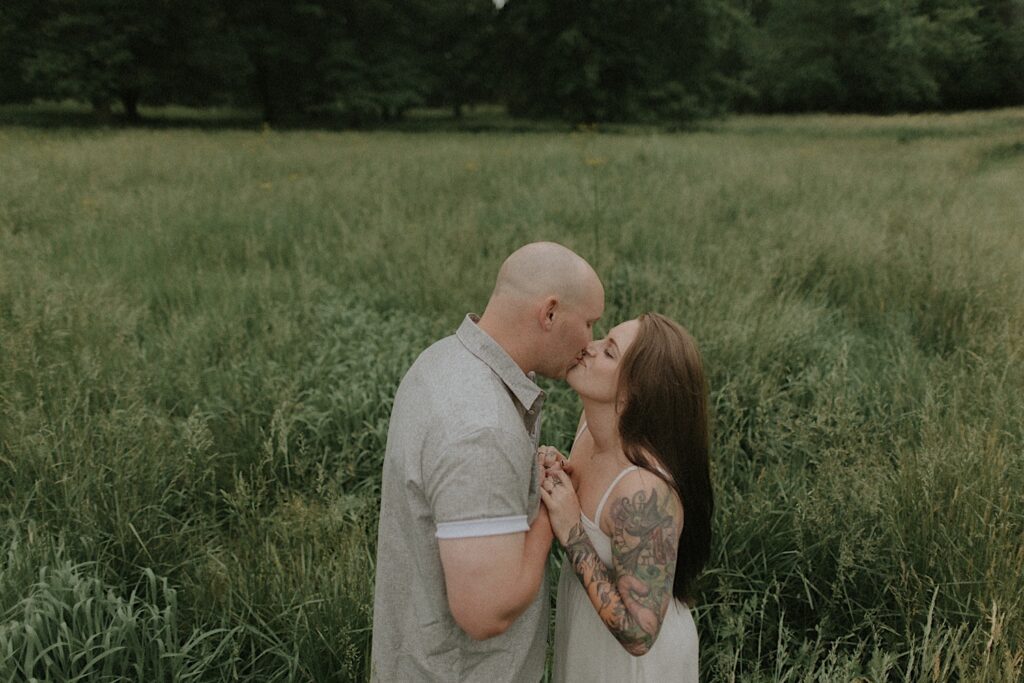 Couple kisses and holds hands. They are standing in a beautiful grassy plain for moody engagement portraits in Lincoln, Illinois. 