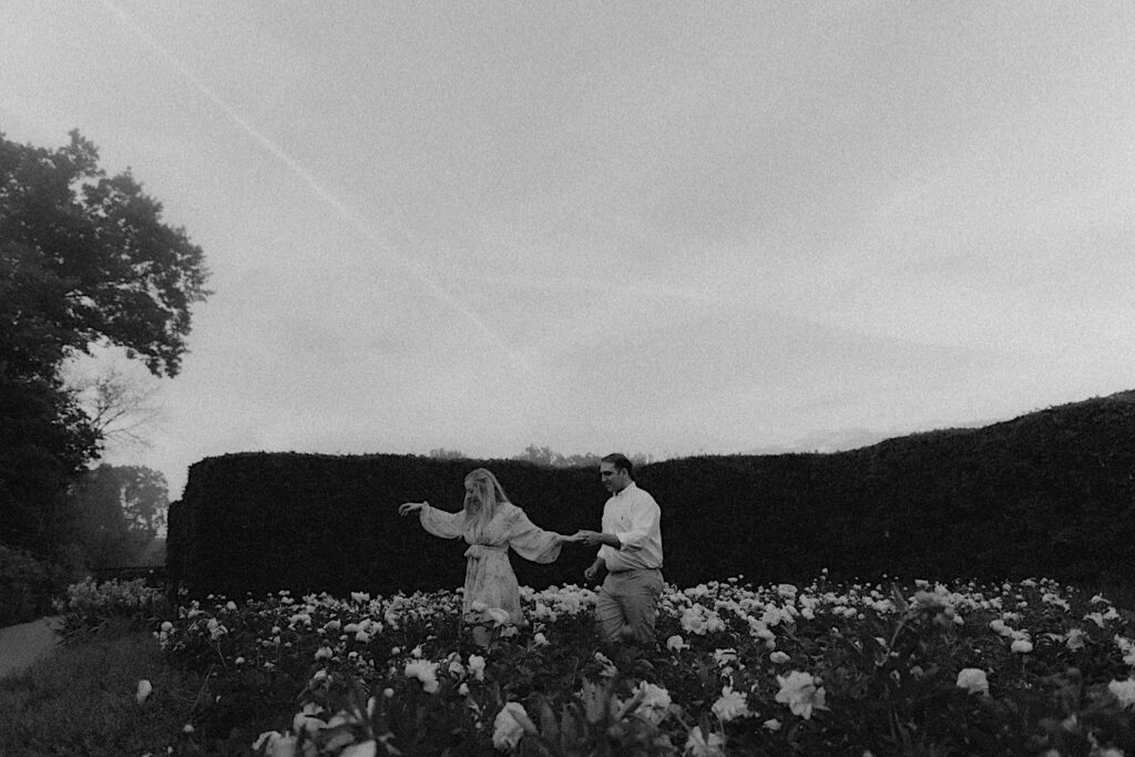 Black and white portrait of couple walking through bushes of beautiful peonies in May during engagement portrait session.