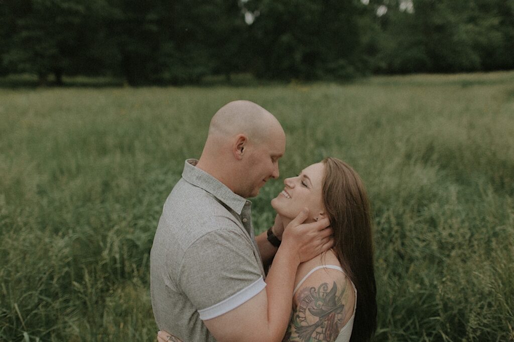 Man holds his fianceé's face as she looks up at him and smiles. They are standing in a beautiful grassy plain for moody engagement portraits in Lincoln, Illinois. 