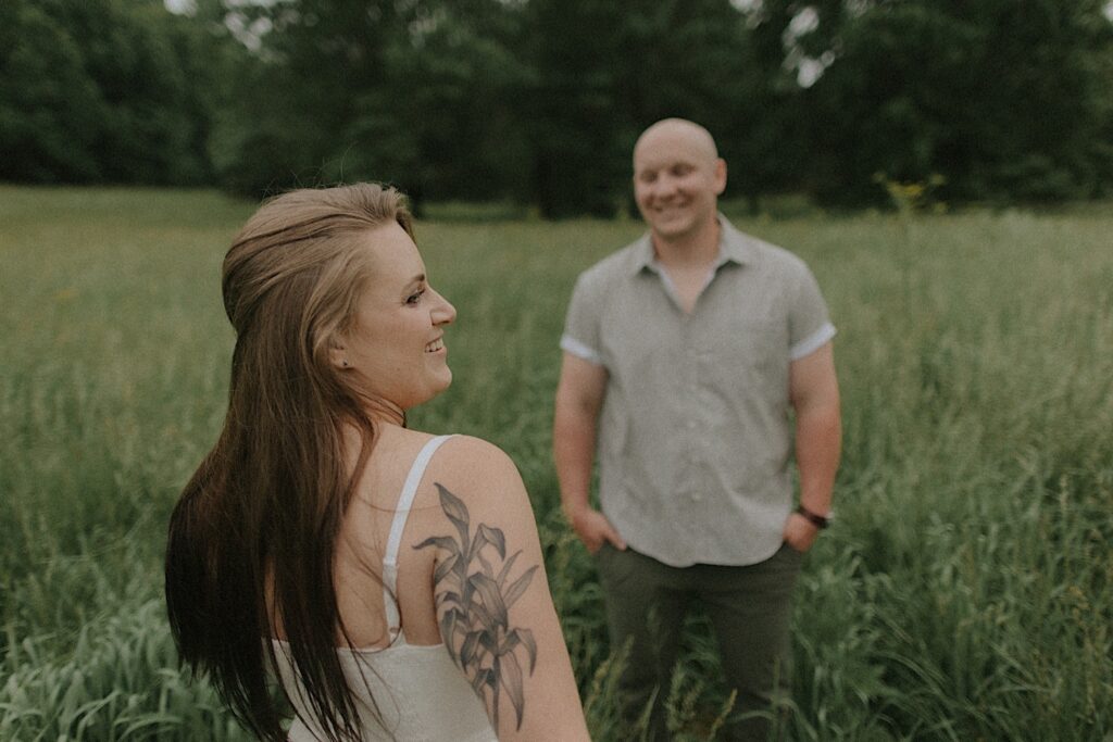 Man looks at a woman as she turns her head to the right side and smiles. They are standing a few feet a part in grassy plains.