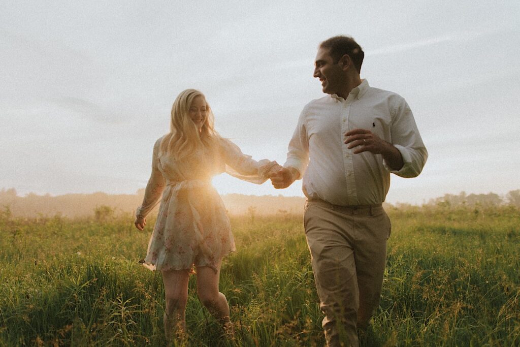 Couple runs through beautiful green field holding hands with golden hour sun shining between them. 