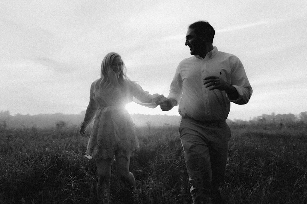 Black and White portrait of couple running through beautiful green field holding hands with golden hour sun shining between them. 