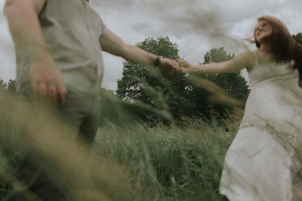 Couple runs through grassy plains with their arms outstretched to hold hands in a moody engagement portrait in Lincoln, Illinois.