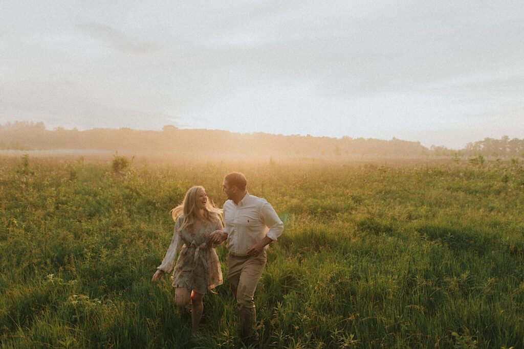 Couple looks at each other while holding hands and running through green field with golden hour sun behind them. 