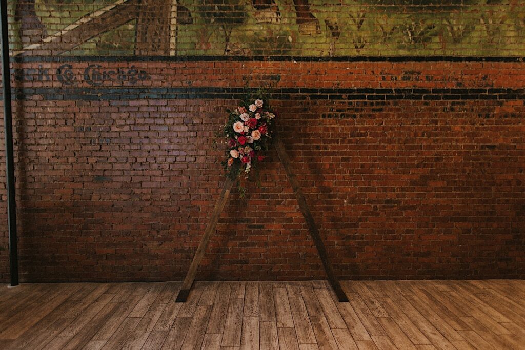 Wooden triangle arch with peach and pink flowers on top of arch in front of brick wall.