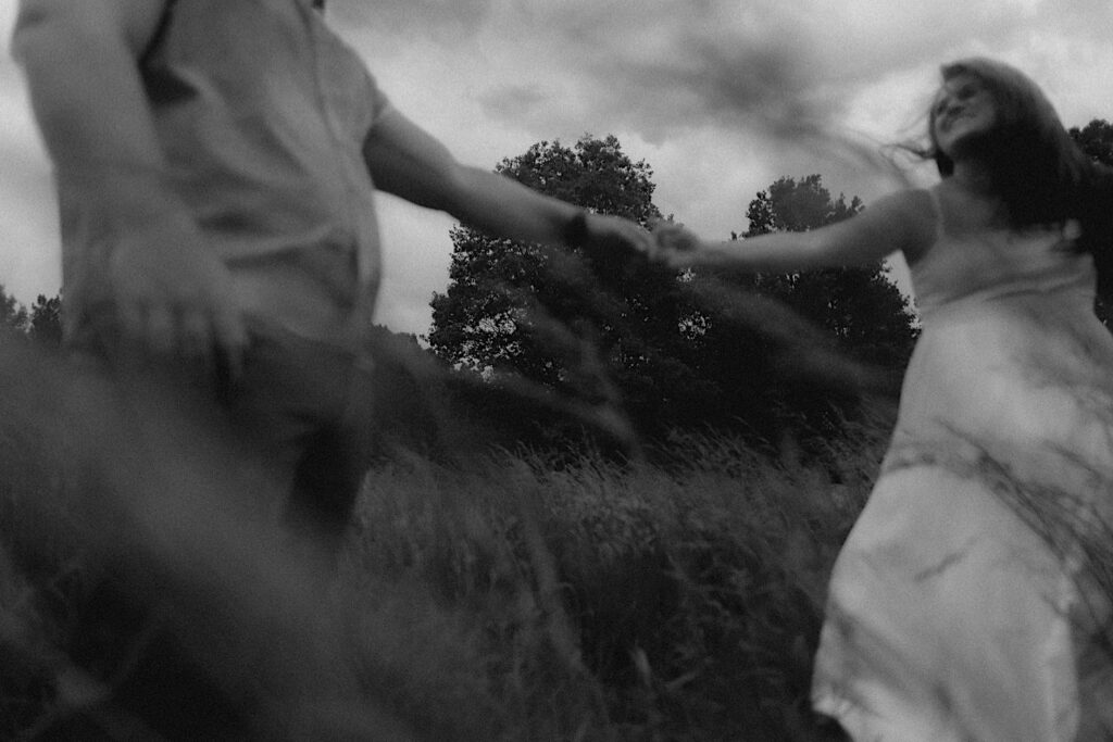 Couple runs through grassy plains with their arms outstretched to hold hands in a moody engagement portrait in Lincoln, Illinois.