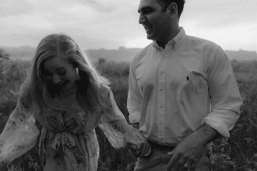 Candid portrait of woman laughing while looking down and holding her fiance's hand as he giggles and they stand in a field. 