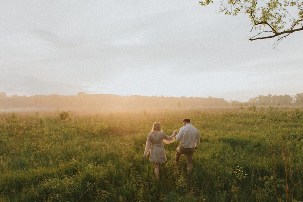 Couple walks with their backs to the camera while walking through grassy field towards golden hour sun setting in the background. 