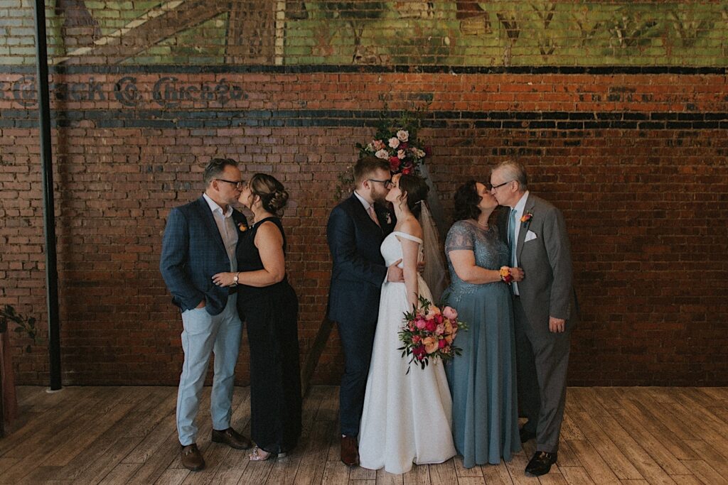 Bride and groom kiss in the center while the Groom's parents kiss to the left of them and the bride's parents kiss to the right of them. They are standing in front of a brick wall in the ceremony space. 