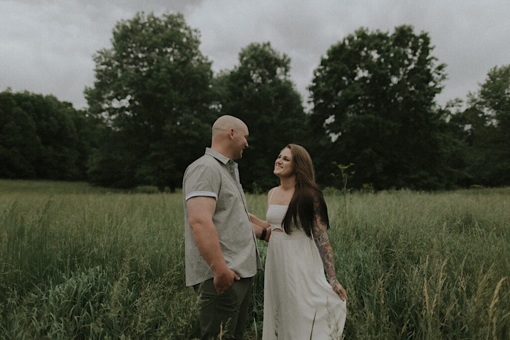 Woman in a white skinny strap dress puts her hands on her fiancé's shoulder as he looks at her smiles. In the background are tall grassy plains in Lincoln, Illinois. 