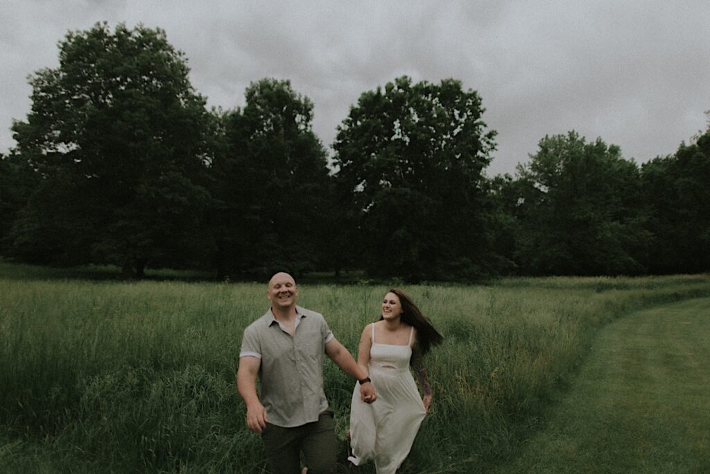 Couple runs through tall grassy plains with trees in the background. 