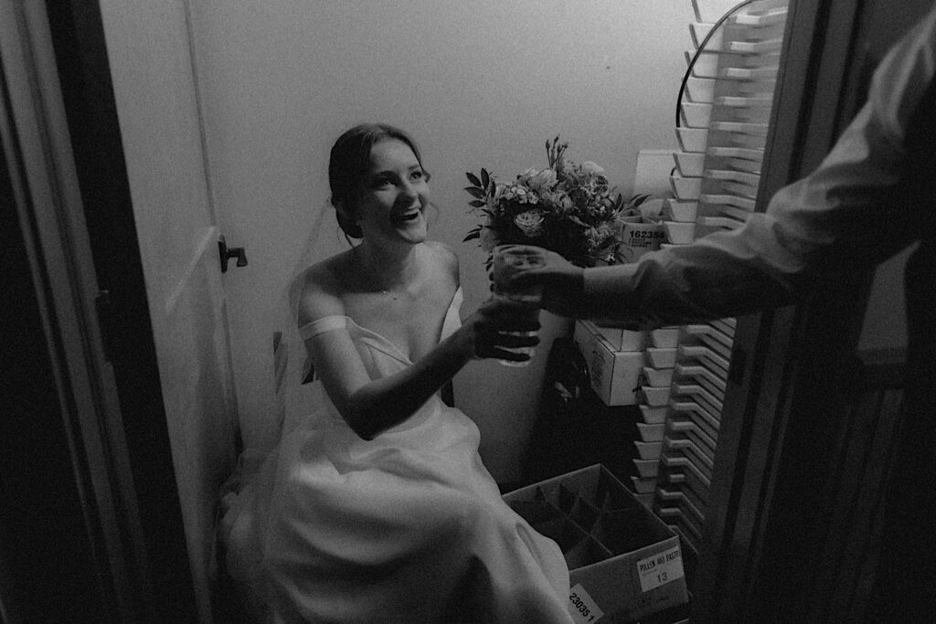 Bride sits on a chair as someone arm reaches into frame and hands her a glass of water while awaiting the wedding ceremony at Reality on Monroe in Central Illinois. 