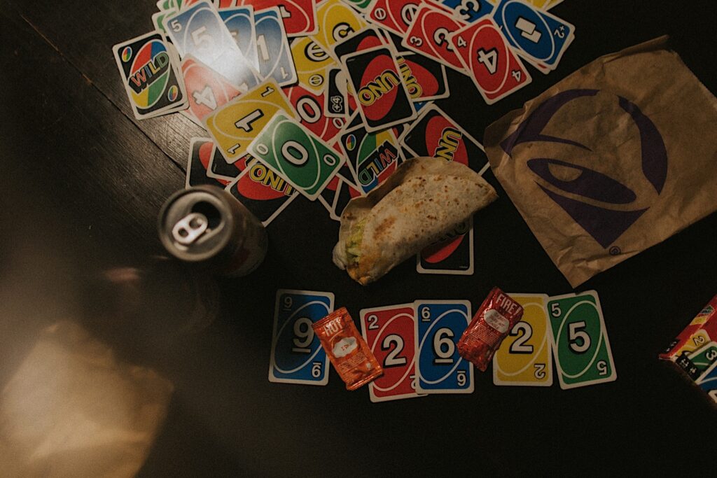 Taco Bell Hot sauce packets sit on-top of Uno cards with a Taco and beer on a table.