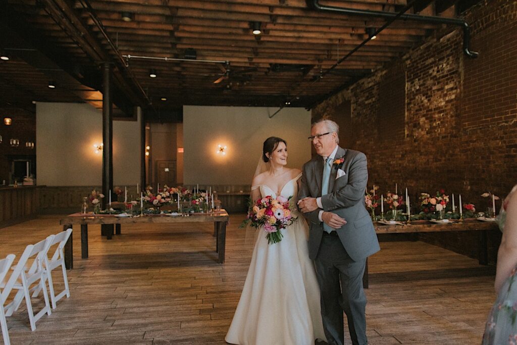 Bride holds onto her father's arm as they walk down the aisle and looks at him. 