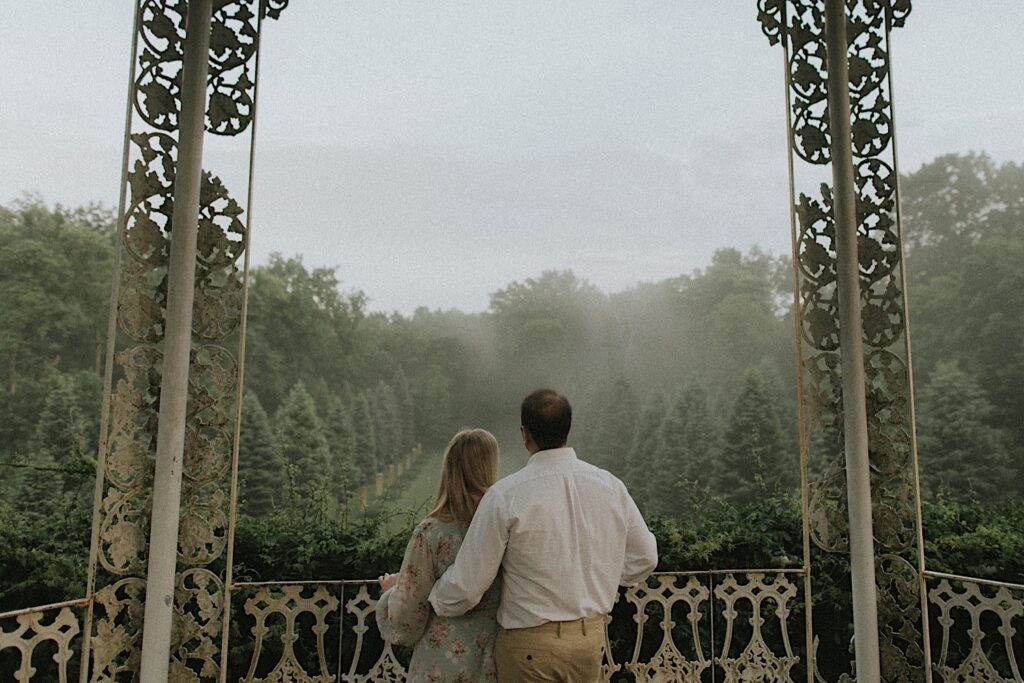 Couple with their backs to the camera look out on balcony of two story gazebo towards large amounts of green trees being hugged by fog. 