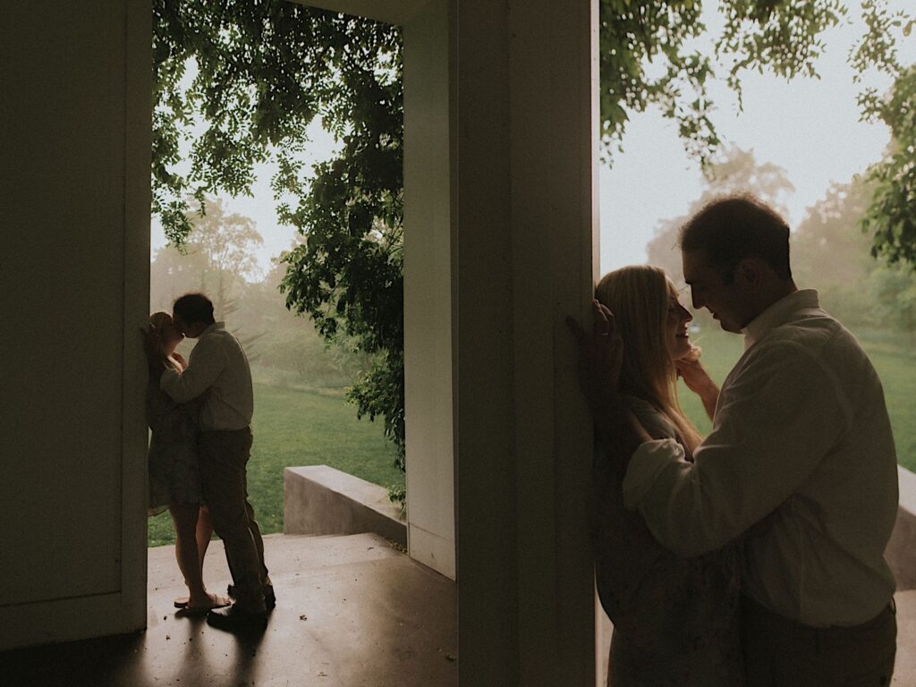 Man kisses his fiance up against a white post romantically with golden hour sun lit behind them. 