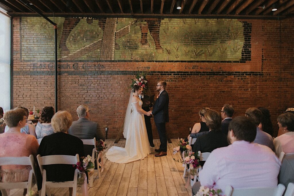 Bride and Groom stand at the end of the aisle and smile at each other holding each other's hand during wedding ceremony at Reality on Monroe. 