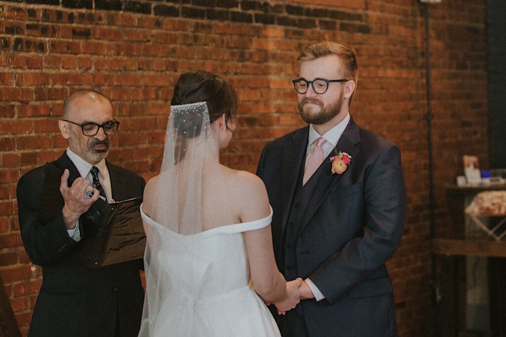 Groom smiles at bride during wedding ceremony in exposed brick room at Reality on Monroe in Central Illinois. 