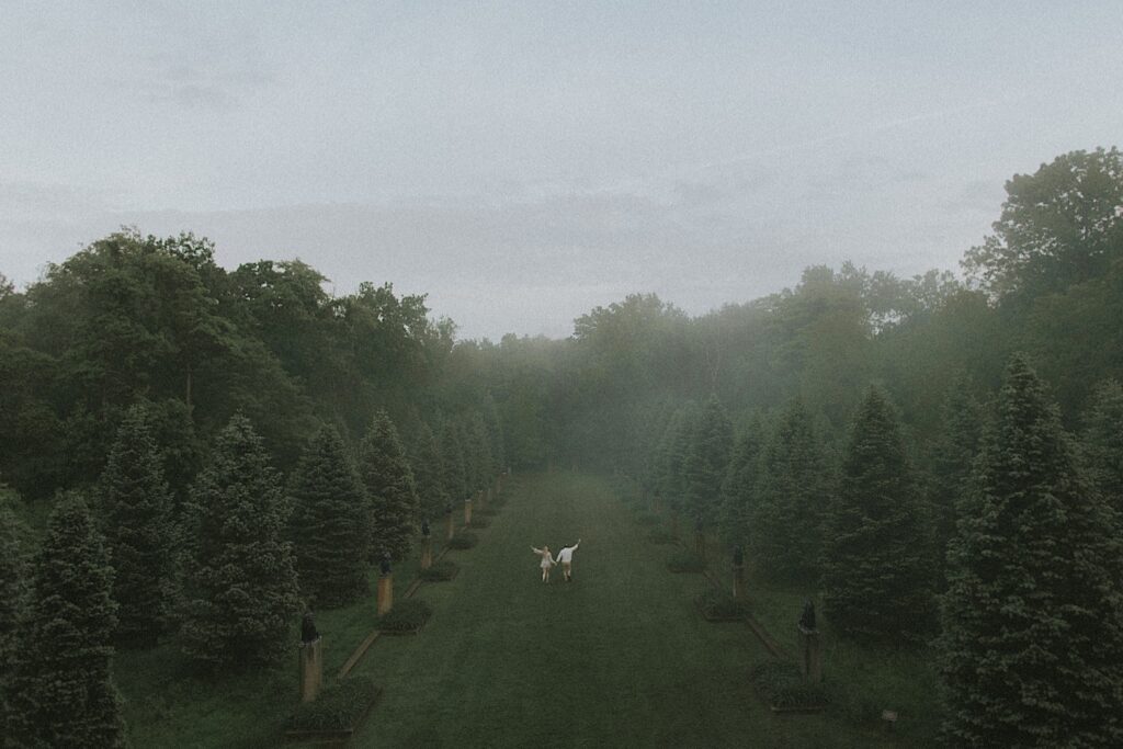 Cinematic portrait of couple running through open grass surrounded on both sides by lines of trees with hazy fog surrounding them. 