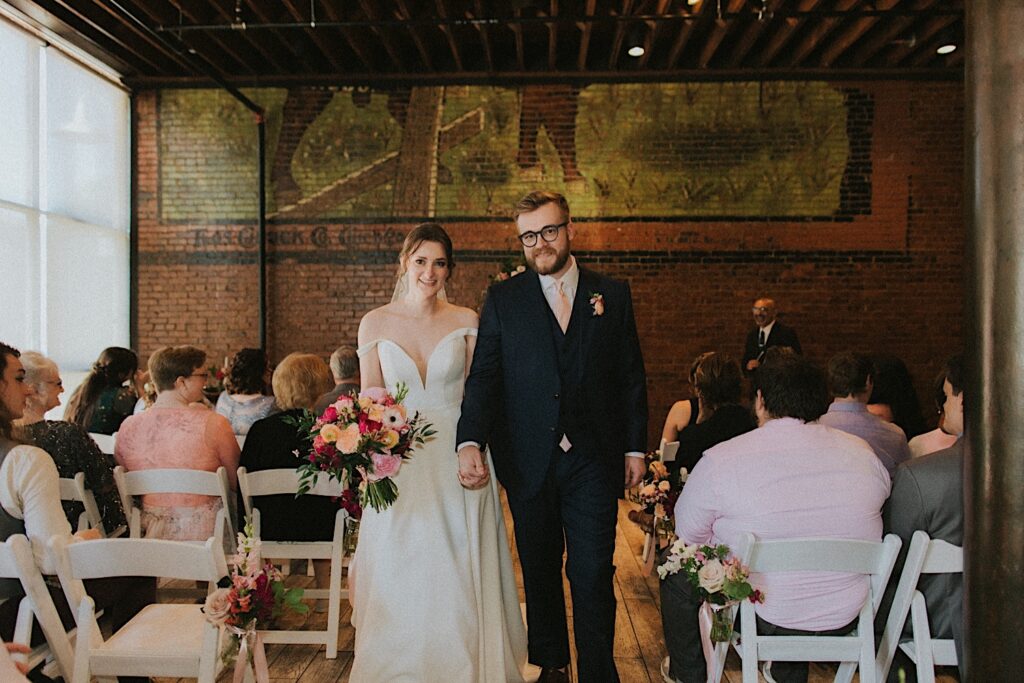 Bride and Groom walk down the aisle after wedding ceremony at smile at the camera as the guests remain seated around them. 