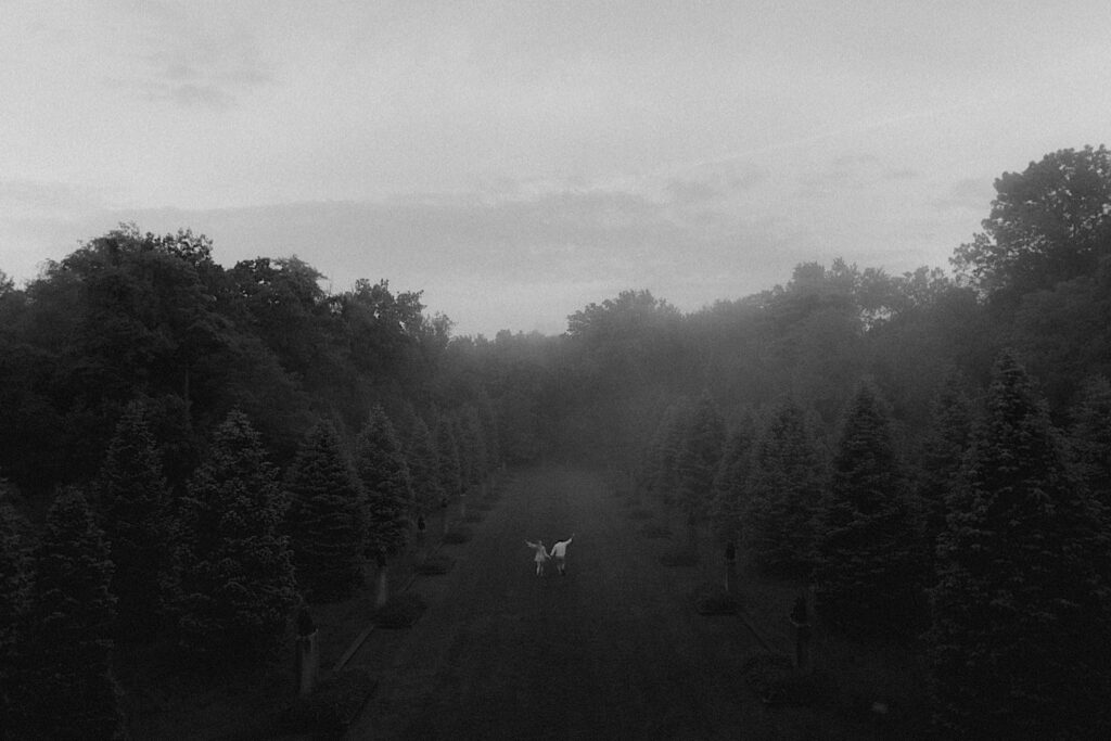 Cinematic portrait of couple running with their hands in the air through open grass surrounded on both sides by lines of trees with hazy fog surrounding them.  