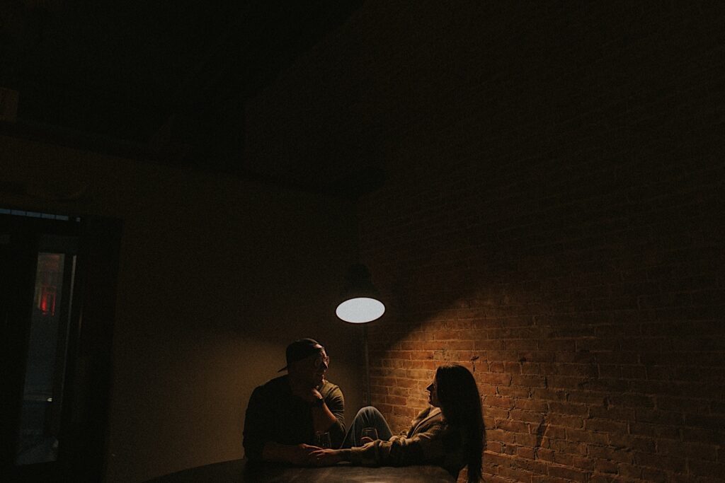 Bride and Groom sit facing each other with a lamp hanging behind them for a moody silhouette portrait. 