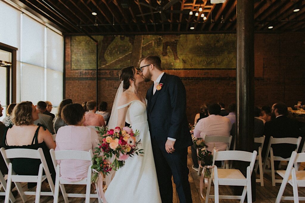 Bride and Groom kiss at the end of the aisle while the guests remain seated around them in beautiful industrial room at Reality on Monroe in Central Illinois. 