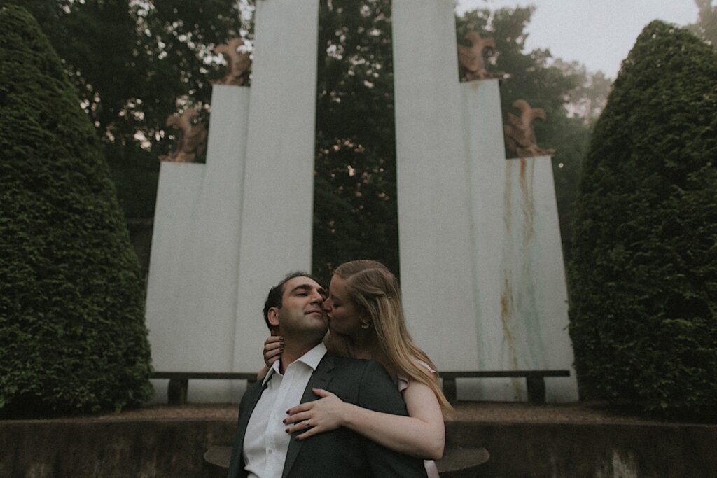 Woman kisses her fiance's cheek from behind in front of white towering sculpture in Allerton Park. 