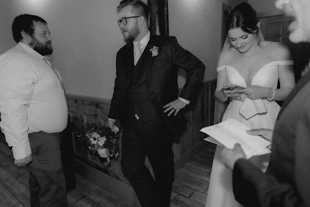 Groom leans up against door frame as bride stands next to him looking at her phone in candid shot by Central Illinois Wedding photographer. 