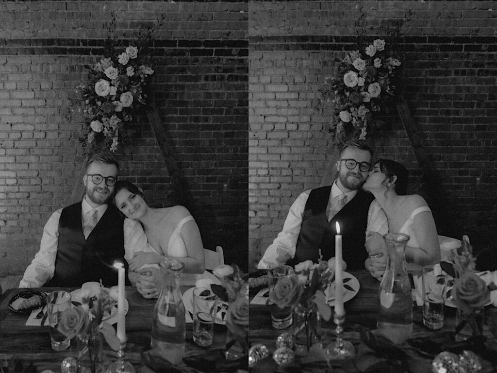 Bride lays her head on groom's shoulders as they sit at the head table during dinner at Reality on Monroe wedding venue. The table is covered in flowers, candles and their plates with food. 