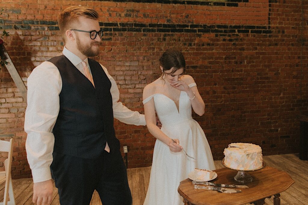 Groom holds onto bride's arm as she sticks her fork into beautiful piece of cut wedding cake during wedding reception at Reality on Monroe.